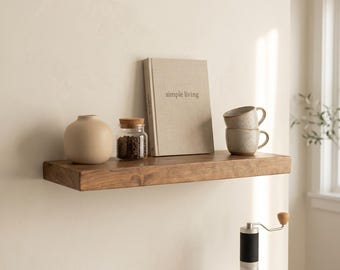 May include: A rectangular wooden floating shelf mounted on a wall, holding a beige ceramic vase, a glass jar with a cork lid filled with coffee beans, a book titled simple living, and two stacked speckled ceramic mugs with handles.