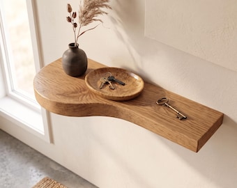 May include: A wall-mounted wooden shelf with a curved edge featuring a dark gray vase with dried grass, a small wooden bowl containing two metal keys, and a single antique-style metal key resting on the surface of the light brown wood grain.