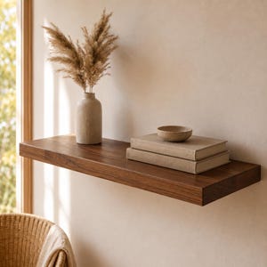 Op de afbeelding: Een rechthoekige, donkerbruine houten zwevende plank aan de muur. Op de plank staat een beige keramische vaas gevuld met gedroogd pampasgras, twee gestapelde beige boeken met harde kaft en een kleine, ondiepe beige keramische kom.