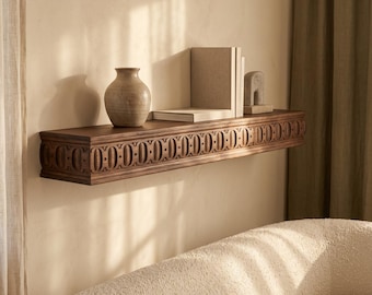 May include: A dark brown wooden floating shelf featuring a repeating carved circular pattern along the front edge. The shelf is mounted on a wall and holds a beige ceramic vase, two upright books, and a small stone sculpture.