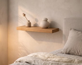 May include: A light-colored wooden floating shelf with two decorative ceramic vases and a small vase with a dried flower. The shelf is mounted on a neutral-toned wall above a bed with a linen duvet.