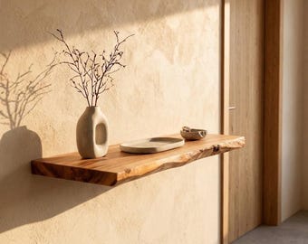 May include: A floating wall shelf made of natural wood with a live edge, featuring a light beige ceramic vase with thin branches, a flat oval ceramic dish, and a small textured bowl arranged on its surface against a textured wall.