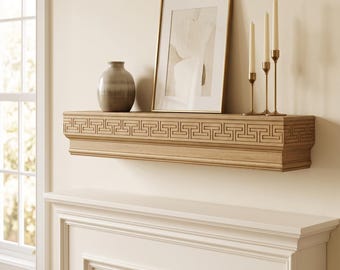 May include: A light brown wooden fireplace mantel shelf featuring a carved Greek key pattern along the front edge. The shelf is mounted horizontally on a wall and supports a small ceramic vase, a framed abstract print, and three tall candles.