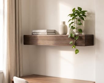 May include: A dark brown wooden floating shelf with a horizontal ribbed texture. On the shelf are two stacked books titled Modernist Homes and The Art of Space, a matte beige ceramic vase, and a trailing green pothos plant in a beige pot.