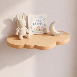 May include: A light-colored wooden wall shelf shaped like a cloud. On the shelf sits a beige knitted rabbit toy, a book titled Little Dreamer by Anna Lee with a pastel illustration, and a smooth, white, crescent moon-shaped decorative object.