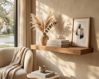 May include: A floating wooden shelf mounted on a beige wall, holding a textured beige vase with dried pampas grass, a stack of three books, and a framed abstract black and white print. Below sits a small side table with a ceramic mug and a book.