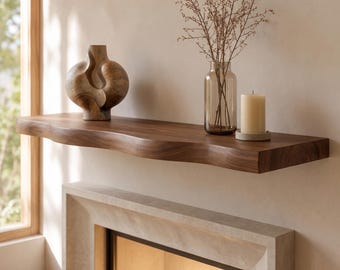 May include: A dark brown wooden floating shelf with a natural live edge, mounted above a fireplace. On the shelf sits a brown abstract wooden sculpture, a clear glass vase containing thin dried branches, and a cream-colored pillar candle on a tray.