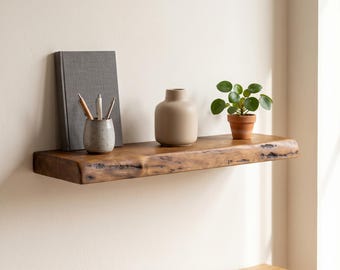 May include: A natural edge wooden floating shelf mounted on a wall, holding a gray book, a speckled ceramic cup with pens, a beige vase, and a small green potted plant. The shelf features a dark brown finish with visible wood grain and knots.