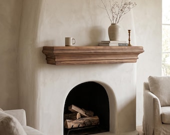 May include: A fireplace with a smooth, off-white plaster surround and a dark wood mantel. The mantel holds a ceramic mug, a stack of books, a textured vase with bare branches, and a brass candlestick. Logs sit inside the arched firebox.
