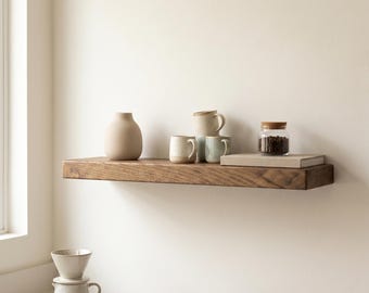 May include: A floating wooden shelf holds a beige vase, three ceramic mugs, a book, and a glass jar with coffee beans. Below, a ceramic pour-over coffee dripper sits next to a small wooden scoop on a white countertop near a window.