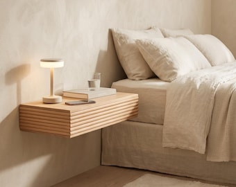 May include: A light-colored wooden floating nightstand with a horizontal ribbed front panel. The nightstand is mounted to a wall and holds a small portable lamp, a closed book, and a smartphone. The surface is smooth and rectangular.