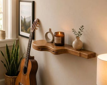 May include: A natural wood wall shelf with an organic, wavy edge profile. The shelf holds a white ceramic ring-shaped vase, a glass jar containing a lit candle with a visible flame, and a small white ceramic vase with a green plant sprig.
