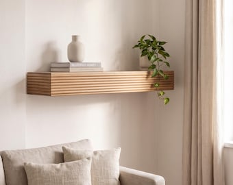 May include: A light-colored wooden floating shelf featuring a horizontal ribbed texture along the front edge. The shelf is mounted on a wall and holds a beige ceramic vase, a stack of books titled Modernist Homes, and a trailing green plant.