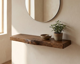 May include: A rectangular floating shelf made of dark brown wood with a natural live edge, mounted on a wall. On the shelf sits a small, round, light-colored ceramic bowl containing keys and a small green succulent plant in a gray ceramic pot.