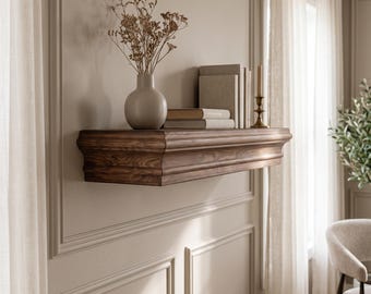 May include: A dark brown wooden floating shelf with a decorative molded edge. The shelf holds a light gray ceramic vase containing dried flowers, a stack of three neutral-colored books, and a thin brass candlestick with a taper candle.