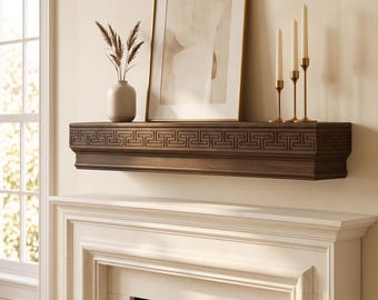 May include: A dark brown wooden fireplace mantel featuring a carved Greek key pattern along the front edge. The mantel is mounted above a white fireplace and supports a framed abstract print, a small beige vase with dried grass, and three gold candle holders.