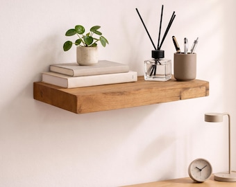May include: A rectangular floating wall shelf made of natural light brown wood with a visible grain pattern. The shelf holds two stacked books, a small potted green plant, a glass reed diffuser with black sticks, and a cylindrical pen holder.