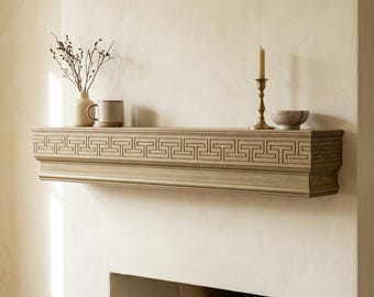 May include: A light brown wooden fireplace mantel featuring a carved Greek key pattern along the front edge. The mantel is mounted on a cream-colored wall above a fireplace opening, with a vase, mug, candle holder, and small bowl on top.