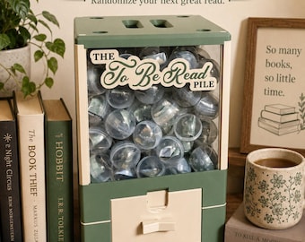 May include: A sage green and cream-colored vending machine labeled The To Be Read Pile, filled with clear plastic capsules. The machine features a dispensing handle and a collection tray, designed to hold small slips of paper for book selection.