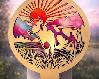 May include: Wooden night light featuring a cow silhouette with colorful accents of a sunset and landscape. The cow has purple and pink spots. The night light is designed to plug into a standard 120V outlet.