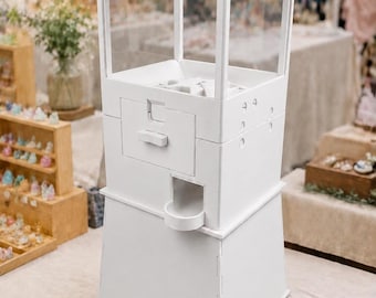 May include: A white vending machine with a clear rectangular top section, a central dispensing mechanism with a small handle, and a lower collection tray. The structure is made of solid white panels with visible seams and a square base design.