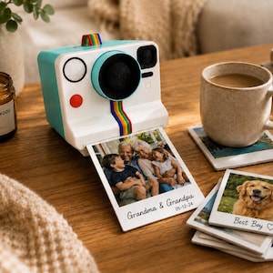 May include: A retro-style instant camera in white and teal with a rainbow stripe detail, printing photos. Beside it, a lit candle, a mug of coffee, and photo coasters with family and dog images. A cozy, lifestyle scene.