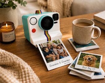May include: A retro-style instant camera in white and teal with a rainbow stripe detail, printing photos. Beside it, a lit candle, a mug of coffee, and photo coasters with family and dog images. A cozy, lifestyle scene.