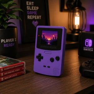 Op de afbeelding: Een paarse handheld gameconsole met een scherm dat een gepixeld zonsondergang boven een stad weergeeft. Het apparaat heeft een D-pad, knoppen en de tekst "GAMES BOOK". Game cartridges en een Nintendo Switch console zijn ook zichtbaar.