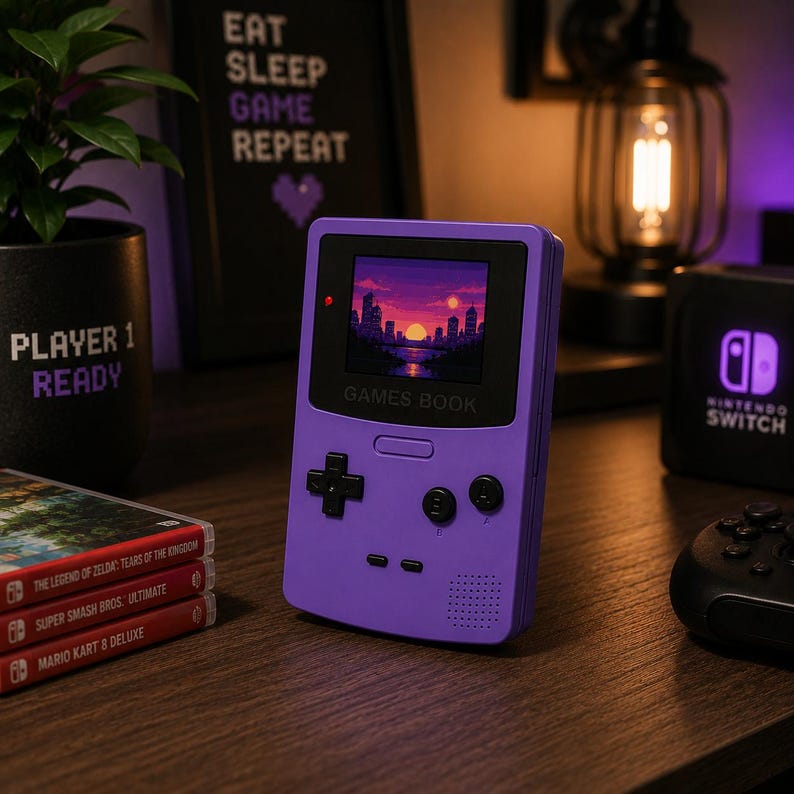 Op de afbeelding: Een paarse handheld gameconsole met een scherm dat een gepixeld zonsondergang boven een stad weergeeft. Het apparaat heeft een D-pad, knoppen en de tekst "GAMES BOOK". Game cartridges en een Nintendo Switch console zijn ook zichtbaar.