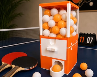 May include: A vertical orange and white plastic dispenser filled with orange and white ping pong balls. The unit features a clear upper storage section, a central white drawer with a handle, and a lower dispensing tray holding one orange ball.
