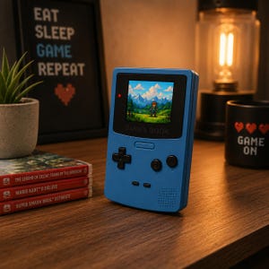 Op de afbeelding: Een blauw draagbaar gamingapparaat met een scherm waarop een videogame-sc&egrave;ne wordt weergegeven. Het apparaat heeft een D-pad en knoppen. De tekst "GAMES BOOK" is zichtbaar. Een ingelijst bord leest "EAT SLEEP GAME REPEAT". Gamecartridges zijn in de buurt opgestapeld.