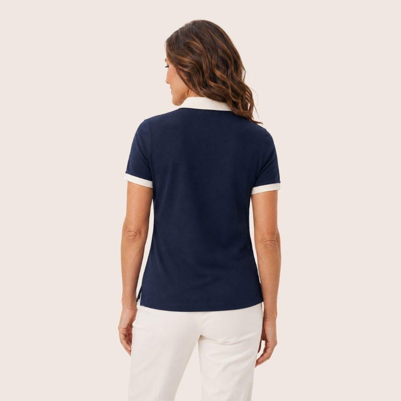 May include: A short-sleeved navy blue polo shirt featuring a white collar and white trim on the sleeve cuffs. The shirt is paired with white trousers. The view shows the back of the garment, highlighting the solid dark blue fabric and contrast details.