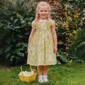 May include: A young girl wearing a yellow dress with a white collar and a floral pattern. The dress has puffed sleeves and a full skirt. She is standing in a grassy area with a basket of colorful Easter eggs in front of her.