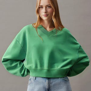 May include: A vibrant green sweatshirt with a V-neck design. The sweatshirt has long sleeves and a cropped fit, with a ribbed hem. The fabric appears soft and comfortable. The model is wearing blue jeans.