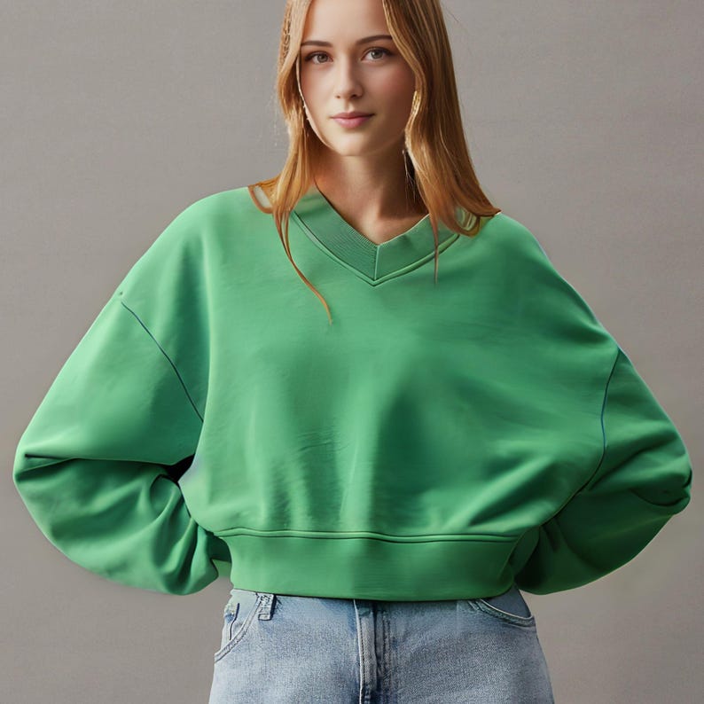 May include: A vibrant green sweatshirt with a V-neck design. The sweatshirt has long sleeves and a cropped fit, with a ribbed hem. The fabric appears soft and comfortable. The model is wearing blue jeans.
