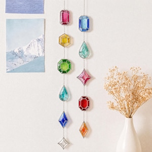 May include: Two vertical hanging decorations featuring multi-colored stained glass shapes in various geometric forms including rectangles, hexagons, diamonds, and teardrops. Each piece is connected by a thin silver metal chain, creating a vibrant display of red, yellow, green, blue, and orange hues.