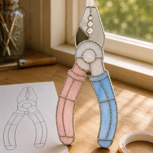 Puede incluir: Una pieza de arte en vitral con forma de alicates, que presenta una sección superior plateada reflectante, un centro blanco y dos mangos en rosa pastel y azul claro, todo unido por finas líneas de soldadura de metal oscuro en un diseño decorativo.