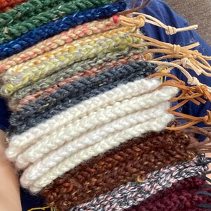 May include: A collection of colorful, hand-knitted wristbands in various shades, including green, blue, white, and brown. Each wristband has a leather cord attached. The knitted texture is visible, showcasing the craftsmanship.
