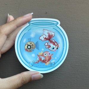 Puede incluir: Una pegatina de pecera azul y blanca con tres peces de dibujos animados dentro. Los peces son un pez marrón y blanco con un ojo negro, un pez rojo y blanco con un ojo rosa y un pez rojo y naranja con un ojo rojo.