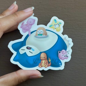 May include: A blue and white sticker featuring a cartoon Snorlax sleeping with other Pokemon characters on top of it. The characters include a pink Jigglypuff, a brown and yellow Charmander, a brown and green Hoothoot, and a yellow and blue Staryu.
