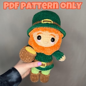 Könnte beinhalten: Ein gehäkeltes Leprechaun-Plüschtier, das einen grünen Hut mit einer goldenen Schnalle trägt und einen Topf mit Gold hält. Der Leprechaun hat einen roten Bart und ein gelbes Gesicht.