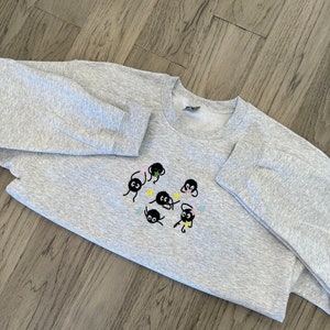 Op de afbeelding: Grijs sweatshirt met een zwart geborduurd ontwerp van meerdere kleine zwarte wezens met gele, blauwe en roze accenten. De wezens zijn in een cirkel gerangschikt.