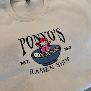 Op de afbeelding: Beige sweatshirt met geborduurd ontwerp. Het ontwerp toont de tekst "PONYO'S RAMEN SHOP" met een stripfiguur die ontspant in een kom ramen. Het ontwerp bevat ook het jaar "2008" en de tekst "EST."