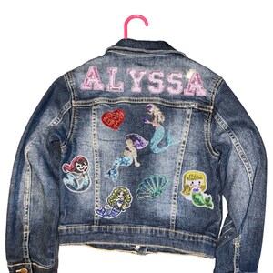 May include: A blue denim jacket with the name "ALYSSA" in pink sequins on the back. The jacket is decorated with several colorful mermaid and seashell patches.