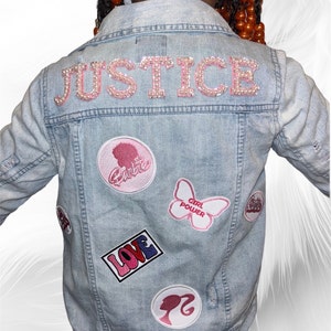 May include: Light blue denim jacket with the word "JUSTICE" spelled out in pink, rhinestone-studded letters across the back. Patches include a Barbie silhouette, a butterfly with "GIRL POWER," and the word "LOVE."