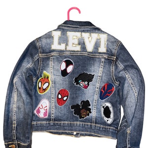 Op de afbeelding: Een blauwe denim jas met de naam "LEVI" geborduurd op de rug in wit met gouden glitter. De jas heeft verschillende kleurrijke patches genaaid op de rug, waaronder Spider-Man-personages.