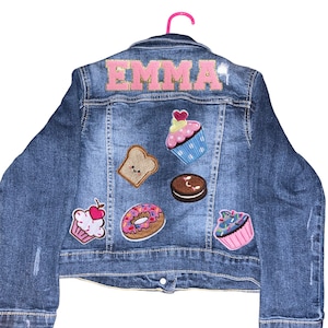 May include: A blue denim jacket with the name "EMMA" embroidered on the back in pink letters. The jacket is decorated with various embroidered patches of cupcakes, donuts, and a slice of bread.