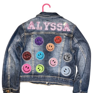 May include: A blue denim jacket with the name "ALYSSA" in pink sequins on the back. The jacket is decorated with colourful smiley face patches.