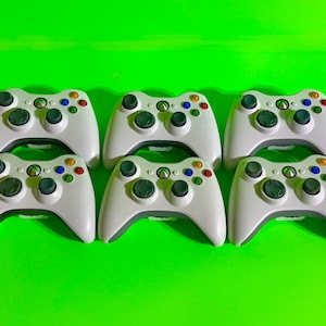 May include: Six white Xbox 360 controllers arranged in two rows of three on a bright green background. Each controller has a gray base, black thumbsticks, and colored buttons.