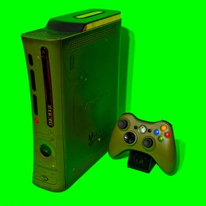 Könnte beinhalten: Grüne Xbox 360 Konsole und passender Controller mit Halo 3 Design. Konsole und Controller sind olivgrün. Der Controller steht auf einem schwarzen Ständer, vor einem leuchtend grünen Hintergrund.